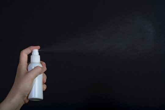 Female hand holding white sprey bottle and spray on dark background 