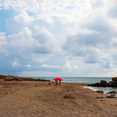 Fototapeta na wymiar Familia debajo de una sombrilla roja en la playa de Mojacar en un dia con nubes