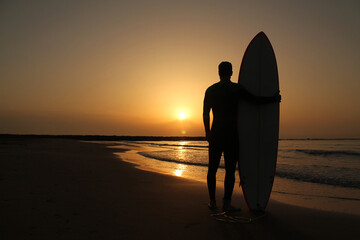 Unrecognizable man is standing with his surfboard seeing a beautiful sunrise on the beach. Copy...