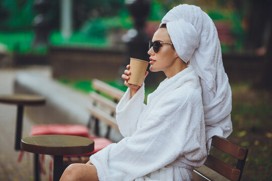 A girl in a robe and a towel on her head sits drinking coffee