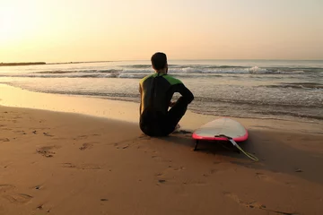 Gordijnen Surfer resting on the shore while sunrise. Thoughtful man relaxes watching the sea after surfing. © ATARES Design