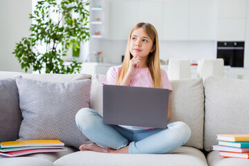 Full length photo of serious minded small pupil girl sit divan legs crossed study remote use laptop think lesson project touch hands chin in house indoors