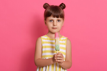 Girl with water ice cream posing isolated over pink background, wearing summer dress with white and yellow stripes, standing with funny knots, looks at camera.