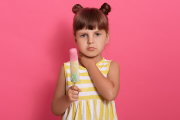 Cute little girl eating ice cream and has sore throat, looking directly at camera with sad facial expression, posing isolated over pink background.