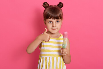 Little happy kid girl holding ice cream and showing thumb up while looking directly at camera, wearing summer dress, has two knots, enjoying tasty sorbet.