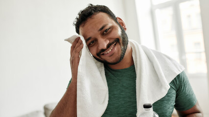 Sweaty from exercising. Portrait of young man smiling at camera, wiping sweat with towel while resting after training workout at home. Sport, healthy lifestyle - obrazy, fototapety, plakaty