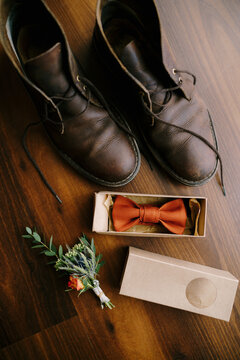 Bow tie in a box for a groom with a boutonniere, and men's boots with untied laces on a brown floor.