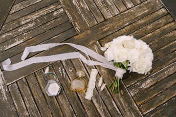 Bouquet of white roses with a long developing ribbon for the bride with garter, perfume bottle, wedding ring, earrings and spikelets on old wooden planks.