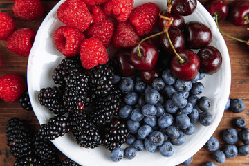 Raspberries, cherries, blackberries, blueberries. View from above. Fresh organic berries in a round plate on a wooden table background.