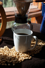 A full coffee cup standing between unroasted coffee beans with rays of sunlight falling down on the beans