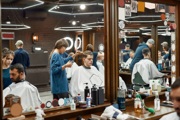 Fototapeta na wymiar Serving client. Professional barber girl, female hairdresser working with hair clipper, serving young handsome guy sitting in barber shop chair