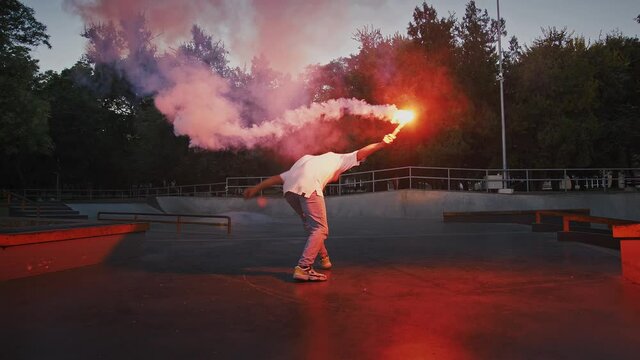 Young man is holding glowing red signal flare, jumping and spinning while performing break dance on pump track of skatepark. Evening, slow motion