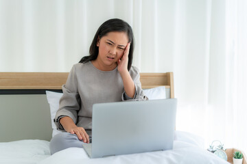 Asian woman in white bed speaking with doctor using tele health technology while sitting on a sofa and headache in bedroom at home while she sick during self quarantine for COVID-19.