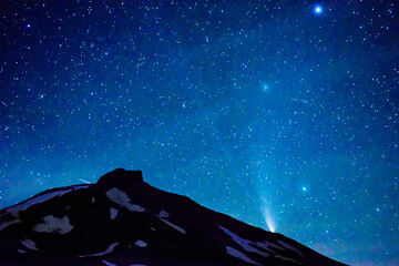 The comet commonly referred to as NEOWISE appears in the night sky over the edge of a partially snow covered mountain peak.