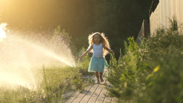 A little girl runs through jets of water to water the lawn in summer. The child is chilling and having fun on a hot summer day near the streams of water.