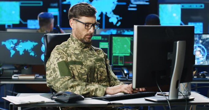 Caucasian male army officer in glasses and uniform sitting at desk, working on computer in analytic secret base in military. Man militarian official typing on keyboard in dark office. Digital support.