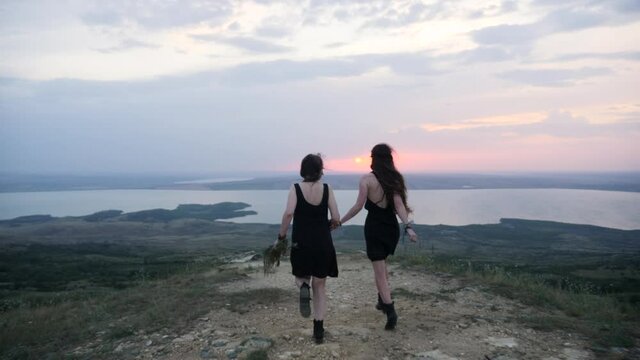 two hippie girls run down the mountainside in black dresses in summer forward to the lake