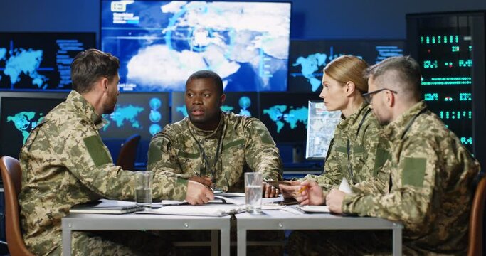 Mixed-races males and females militarian council sitting at table in uniforms, deciding war or defense tactics in dark monitoring room. Multiethnic men and woman officers talking, discussing terrorism