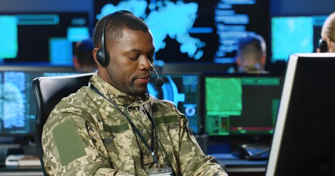 African American male army officer in headset talking and working on computer in analytic secret base in military. Man militarian official typing in dark office. Digital support. Surveillance concept.