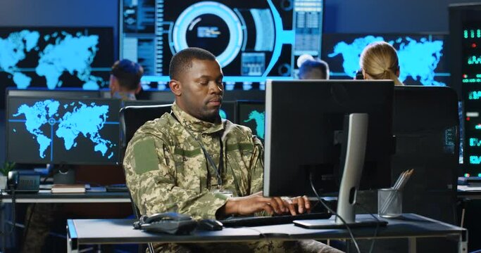 African American male army officer in uniform sitting at desk, working on computer in analytic secret base in military. Man militarian official typing on keyboard in dark office. Digital support.