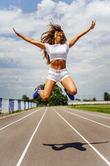 Fototapeta na wymiar Happy young adult caucasian woman on running track - Beautiful female jumping on stadium in sunny summer day - Adult girl jump in air with arms raised - happiness freedom and joy concept