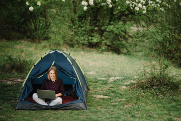 Woman working on laptop in tent in nature. Young freelancer sitting in camp. Relaxing in camping site in forest, meadow. Remote work, outdoor activity in summer. Happy girl relaxing, work on vacation.