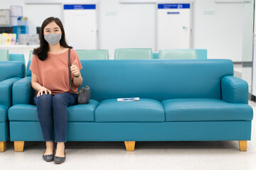 Fototapeta na wymiar Asian woman with hygiene protective face mask over her face sitting on a sofa seat in the hospital with a social distancing protocol while COVID-19 or Coronavirus outbreak. Using face mask concept.