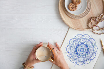Female hands close up draw Decorative round floral mandala. Hobby and home relaxation. A mug of coffee and cookies on a wooden tray. White background.