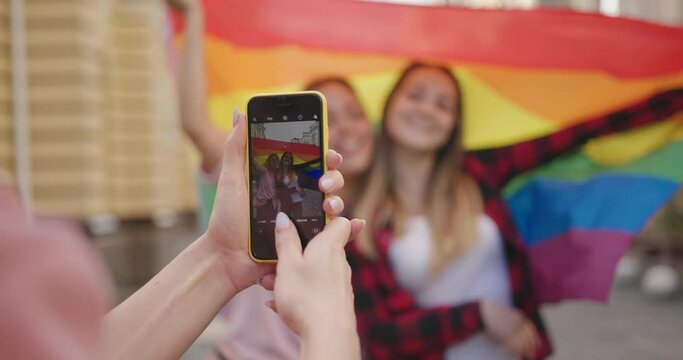 Girlfriend makes photo with smartphone of happy loving homosexual lesbian LGBT couple holding rainbow flag hugging at city street.