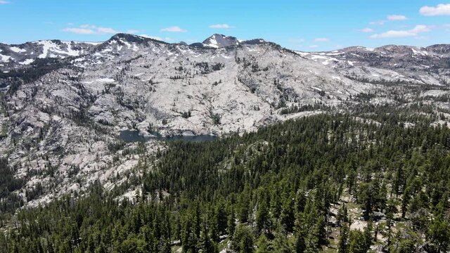 Bird view of Tahoe National Forest during early Summer. Lots of pines right above the image and some snow on the top of the background mountains. The is also a lake in the middle of the footage.