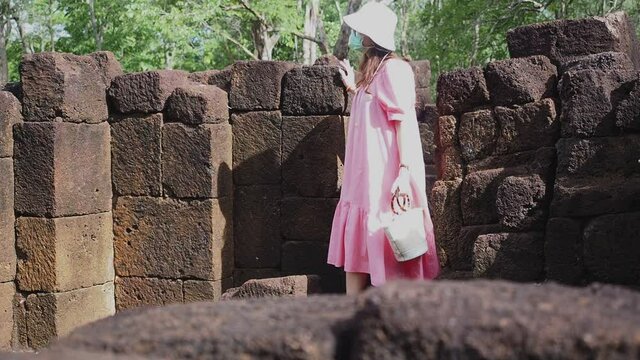 Asian woman tourist in pink dress with medical face mask at "Muang Singha" ancient ruin temple in Kanchanaburi, Thailand. new normal after reopening from covid19 outbreak.