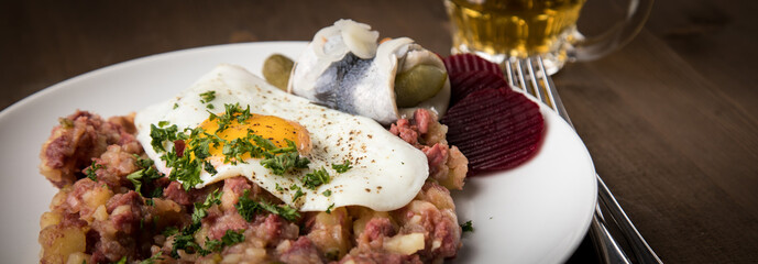 Norddeutscher Hamburger Labskaus mit Rindfleisch Corned Beef, Rote Beete, Kartoffeln, Saure Gurken, Spiegelei, Rollmops und Bier auf Holztisch