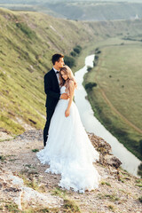 Full length body portrait of young bride and groom enjoying romantic moments outside at sunset in beautiful summer day. Wedding couple. Standing face to face with the green hills on background.