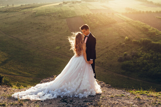 Full length body portrait of young bride and groom enjoying romantic moments outside at sunset in beautiful summer day. Wedding couple. Standing face to face with the green hills on background.