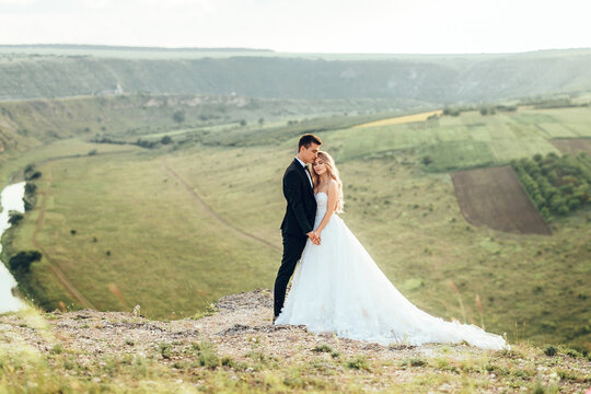 Full length body portrait of young bride and groom enjoying romantic moments outside at sunset in beautiful summer day. Wedding couple. Standing face to face with the green hills on background.