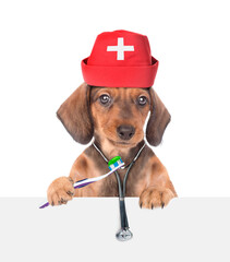 Dachshund puppy wearing doctor's hat and stethoscope holds a toothbrush and looks  above empty white banner. isolated on white background