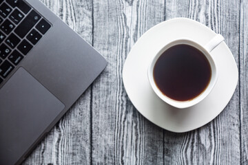 Coffee in a white cup on a white saucer and a laptop on a gray wood background