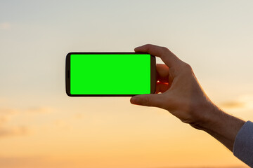 The hand with the phone in a horizontal position on the sunset sky background. Smartphone with a green screen, copy space.