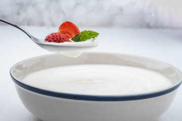 Plain yogurt on a spoon with fresh slice strawberry and mint leaves on top hanging above of plain yogurt in bow.