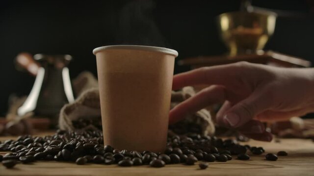 Fingers depicting a man walk to brown paper cup. Hand taking it away. Enjoying tea or coffee to go. Humorous, conceptual, funny acting coffee drinking. Scattered coffee beans, wood desk.