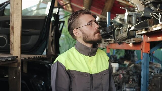 A young bearded service station worker in uniform stands near a shelf with spare parts and chooses the part to repair the car. Close-up