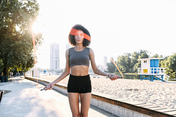 Photo in motion of a jumping girl with a skipping rope. A woman with an Afro hairstyle training outdoor. Midshot