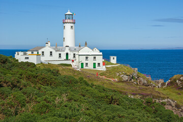 Fototapeta na wymiar The white painted lighthouse at Fanad Head, Donegal, Ireland stands on a cliff top above the blue Atlantic Ocean, a safety beacon for shipping in the dangerous coastal waters around the rocky shores.