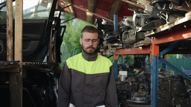 A young bearded auto mechanic with glasses walks between the racks of spare parts and stops with his arms crossed over his chest. Close-up. Slow motion