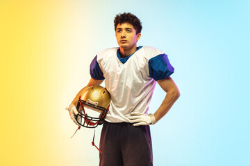 American football player isolated on gradient studio background in neon light. Professional sportsman posing confident and strong during game playing. Concept of sport, movement, achievements.