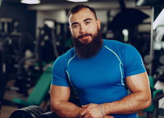 Fototapeta na wymiar Bearded young man bodybuilder in blue t-shirt standing in gym