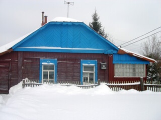 Large snowdrifts. Old painted wooden house in the village. Wooden fence in front of the house. Snow is lying on the roof of the house