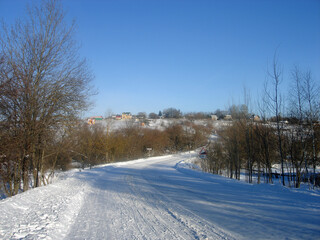 Fototapeta na wymiar Sunny winter day. A winding road covered in snow goes up to the village. Trees with no leaves stand along the road