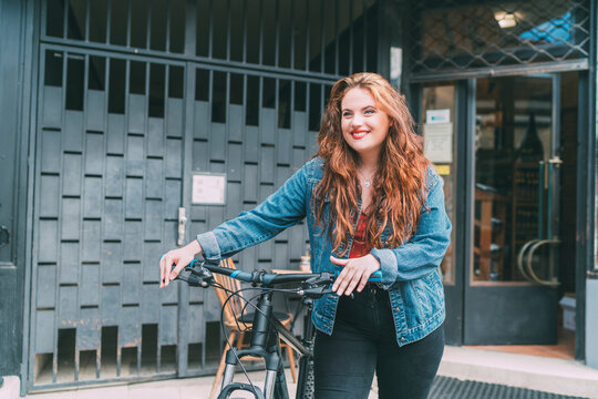 Fashion portrait of smiling Red curled long hair caucasian teen girl on the city street walking with bicycle. Natural people beauty urban life concept image.