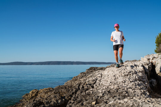 Young female dressed running sporty clothes and pink cap enjoying morning jogging along the rocky calm sea coast. Sporty people trail running activities on vacation time concept image.